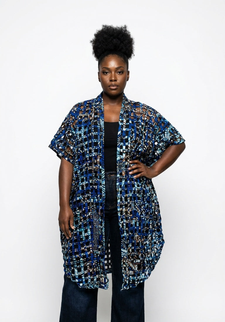 A model stands against a white studio background wearing a short-sleeved, open-front cardigan with a blue, black, and white abstract pattern, paired with dark wide-leg jeans.